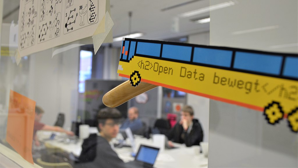 Open Data beweg steht auf einem Bus aus Pappe, der an einem Fenster hängt. Im Hintergrund sitzen Menschen mit Laptops am Tisch.