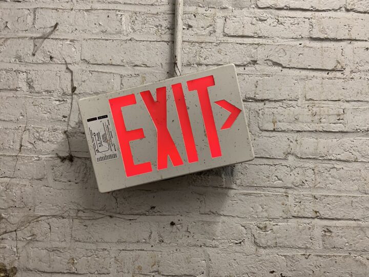 Das Foto zeigt eine weiße Wand aus Backsteinen, an der schief ein weißes Schild mit roter Leuchtschrift hängt. Der Schriftzug lautet Exit, das englische Wort für Ausgang.
