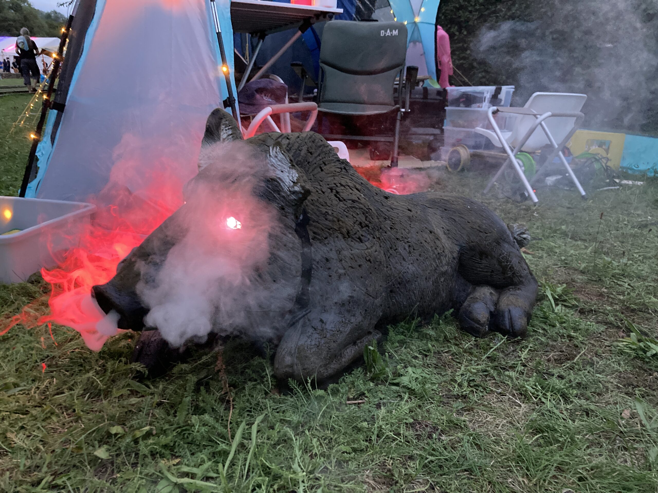 Zu sehen ist eine sehr lebensähnliche Skulptur eines Wilschweins, das vor einem offenen Zelt liegt. Das Schwein dampft aus der Nase und die Augen sind magentarot erleuchtet.