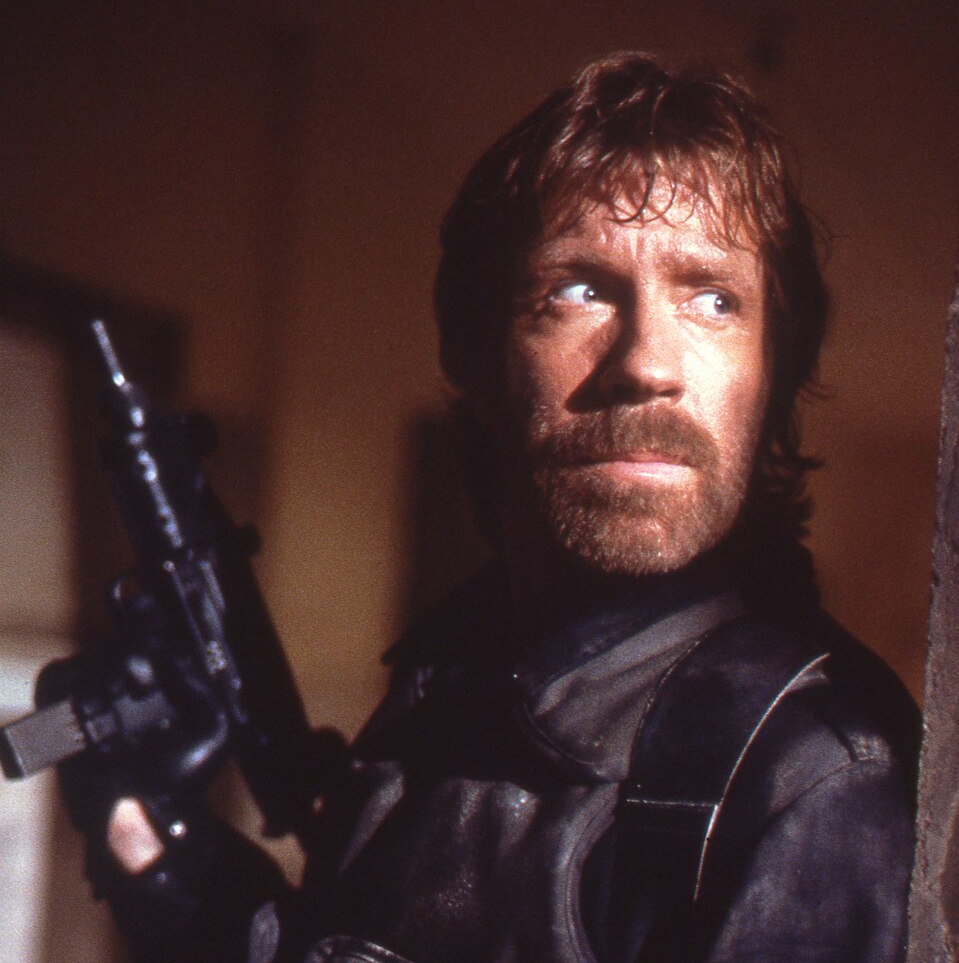 Chuck Norris in schwarzer Kleidung mit einer Waffe in der Hand.