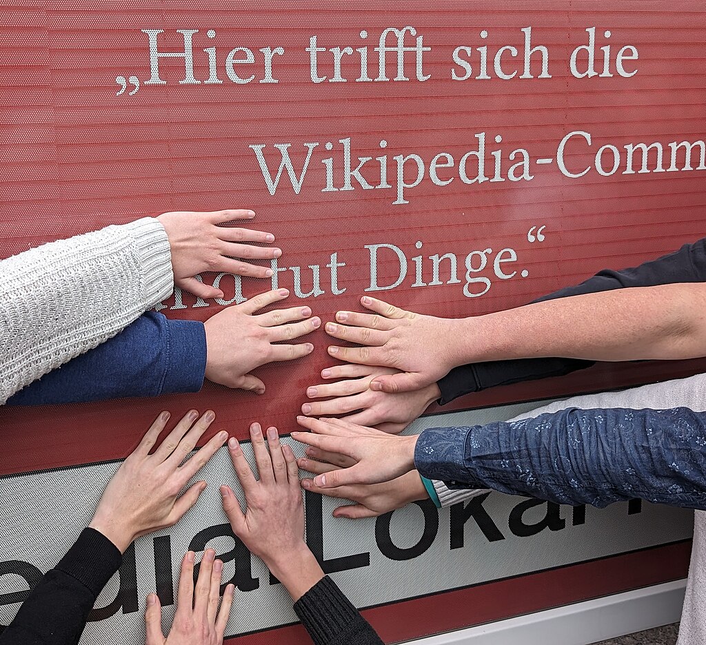 Ein Leitspruch des Lokal K vor rotem Hintergrund: "Hier trifft sich die Wikipedia-Community und tut Dinge." Neun Hände berühren das Poster.
