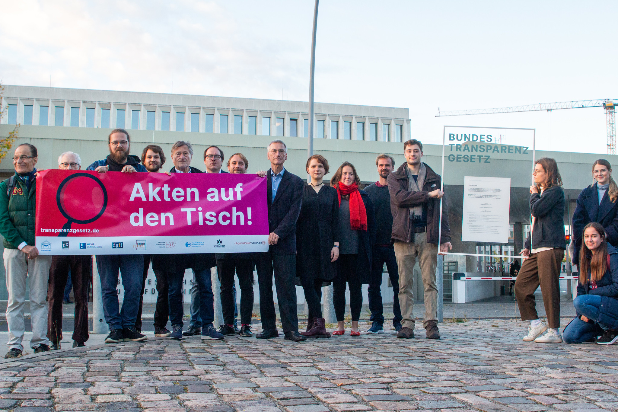 Menschengruppe hält ein Banner in der Hand, darauf steht: "Akten auf den Tisch". Daneben halten zwei Menschen einen Glasrahmen, mit einem Zettel und der Aufschrift: "Bundestransparenzgesetz"