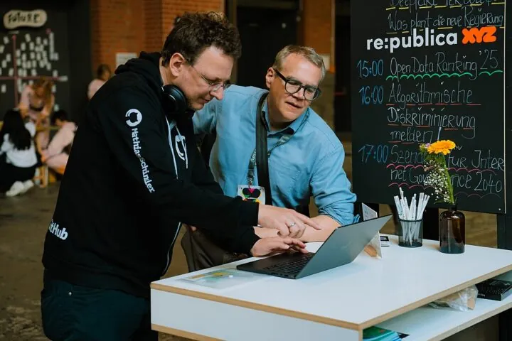 Das Bild zeigt zwei Mit-Gründer des Vereins Wikimedia Deutschland, die bei der re:publica 2025 am Stand vom Bündnis F5 in einen Laptopbildschirm schauen: Open-Data-Experte Mathias Schindler (l.) und Arne Klempert, ehemaliger Geschäftsführer von Wikimedia Deutschland.