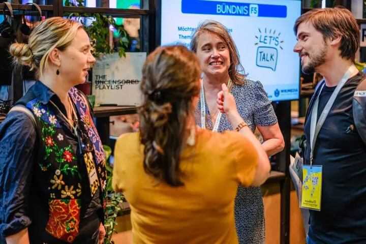 Das foto zeigt drei Frauen und einen Mann, die sich am re:publica Stand von Wikimedia Deutschland und vom Bündnis F5 unterhalten. Im Hintergrund ist ein Monitor zu sehen, auf dem das strahlendblaue Logo vom Bündnis F5 zu sehen ist. Das Foto stammt aus dem Jahr 2023.