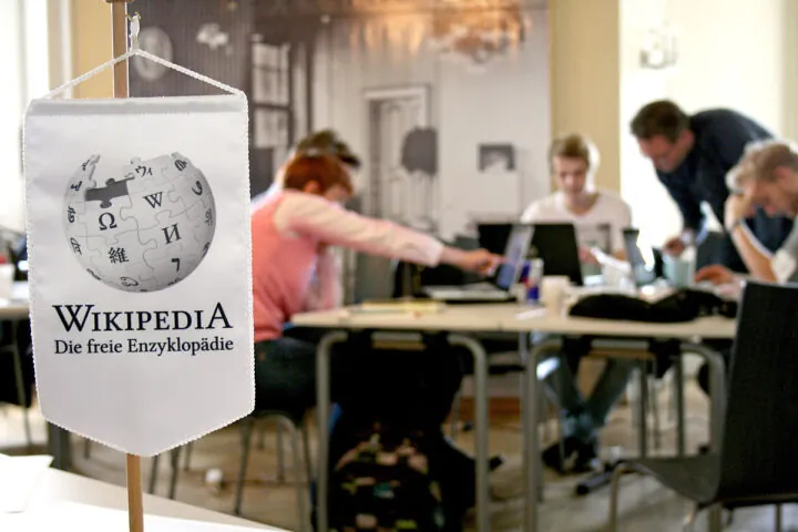 Im Vordergrund ist eine kleine Fahne mit dem Wikipedia-Logo zu sehen. Dahinter sitzen mehrer Menschen vor ihren Laptops.
