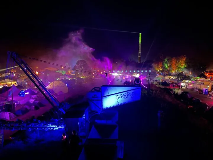 Eine Luftaufnahme vom Gelände des Chaos Communication Camps im Ziegeleipark in Mildenberg, Brandenburg. Es ist Nacht und blaue und lilafarbene Lichter erleuchten die Zelte, alte Ziegeleigebäude und Kunstinstallationen. Auf einem Kaminturm ist ein Scheinwerfer installiert, der die Bahn von Satelliten am Nachthimmel verfolgt.