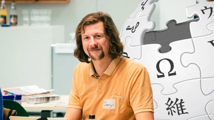 Conny in einem orangen Shirt sitzt an einem Tisch. Im Hintergrund der Wikipedia-Globus.