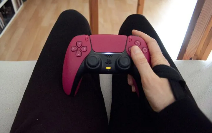Eine sitzende Person hält in der rechten Hand mit Handgelenkbandage einen Playstation 5 Controller.