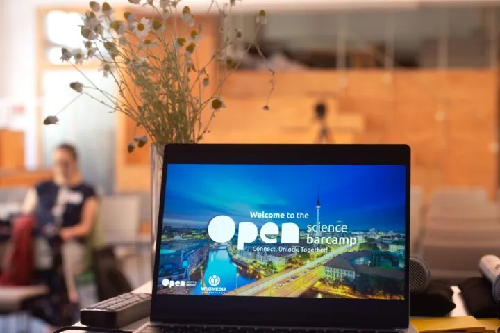 Ein aufgeklappter Laptop zeigt eine Nachtaufnahme von Berlin und die Aufschrift „Welcome to the Open Science Barcamp – Connect, Unlock, Together!